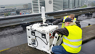 Two technicians in high-visibility vests installing a mobile monitoring unit on a rooftop to assess signal quality and network performance.