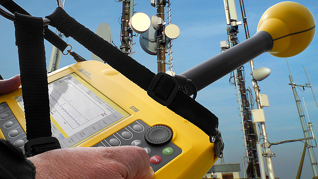 A technician using a yellow handheld spectrum analyzer to test signal quality near a communication tower, representing LS telcom’s acceptance testing services for network validation.